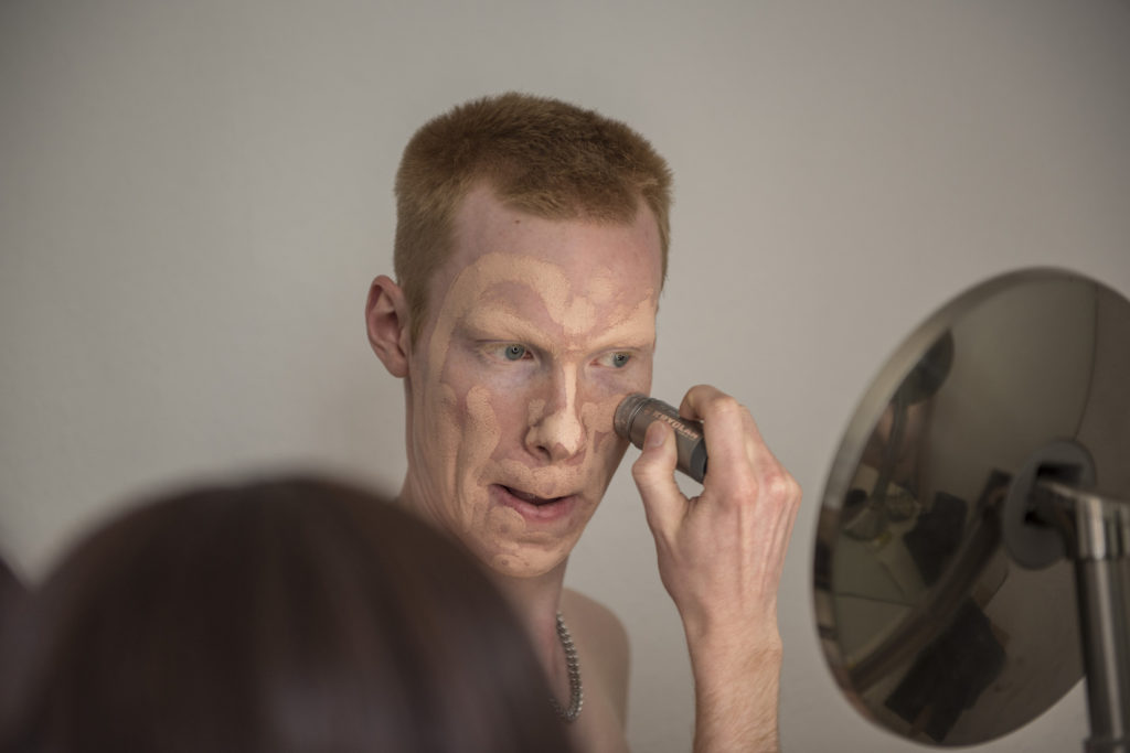 an Kollenbach schaut in den Spiegel und malt mit einem Pinsel Schichten von Make-up in sein Gesicht.